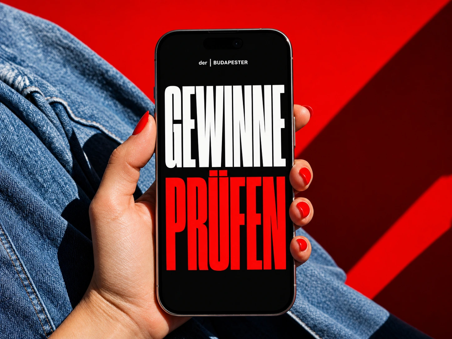 Hand mit roten Fingernägeln hält ein iPhone vor blauer Jeans und rotem Hintergrund. Auf dem schwarzen Display stehen der | BUDAPESTER, GEWINNE in Weiß und PRÜFEN in Rot.