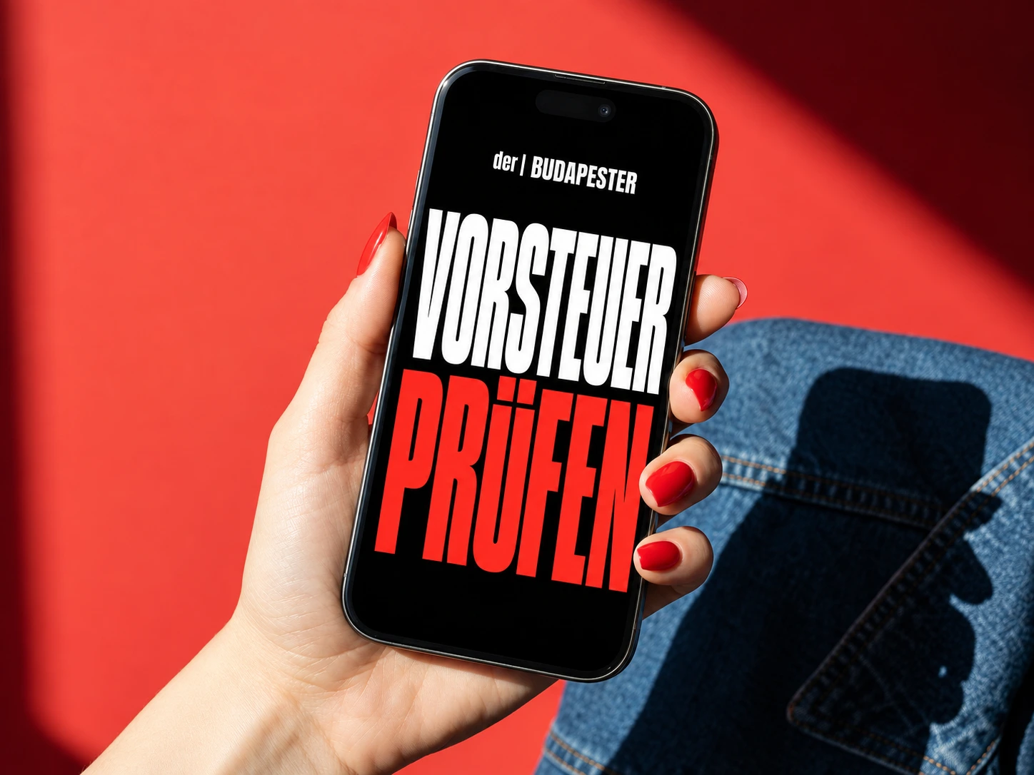 Hand mit roten Fingernägeln hält ein schwarzes iPhone vor rotem Hintergrund und blauer Jeans. Auf dem Display stehen der | BUDAPESTER, VORSTEUER in Weiß und PRÜFEN in Rot.
